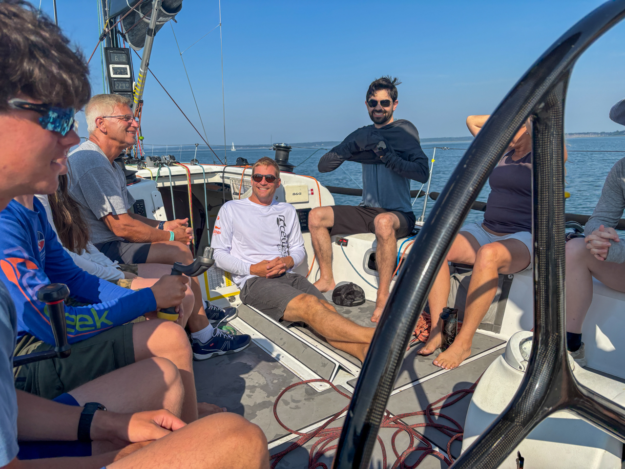 Group of people seated and standing on a sailboat, chatting and smiling on a sunny day at sea with rigging and ropes around them.
