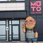 Cartoon mascot outside a pizza shop with pink glasses and overalls, posing with a peace sign next to the 'MOTO ODD PIZZA' sign.