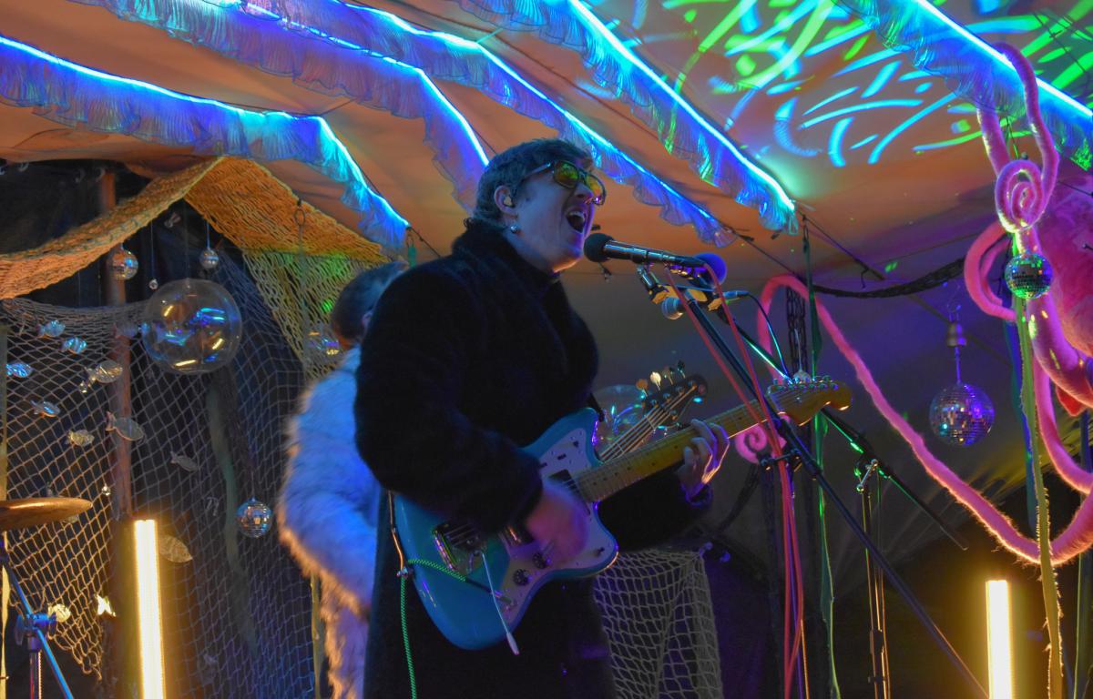 Lead singer and guitarist of a psych-rock band sings into a microphone on an underwater-themed stage.