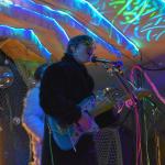 Lead singer and guitarist of a psych-rock band sings into a microphone on an underwater-themed stage.