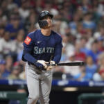 Seattle Mariners batter in navy jersey stands at the plate, gripping a bat with a face-guard helmet.
