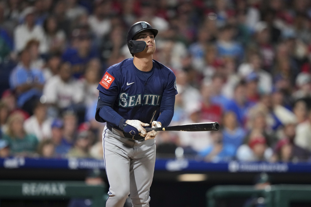 Seattle Mariners batter in navy jersey stands at the plate, gripping a bat with a face-guard helmet.