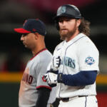 Seattle Mariners batter in white uniform with helmet and 50 Seasons patch stands at the plate, while a Cleveland pitcher/infielder in gray with a red cap lingers behind him.