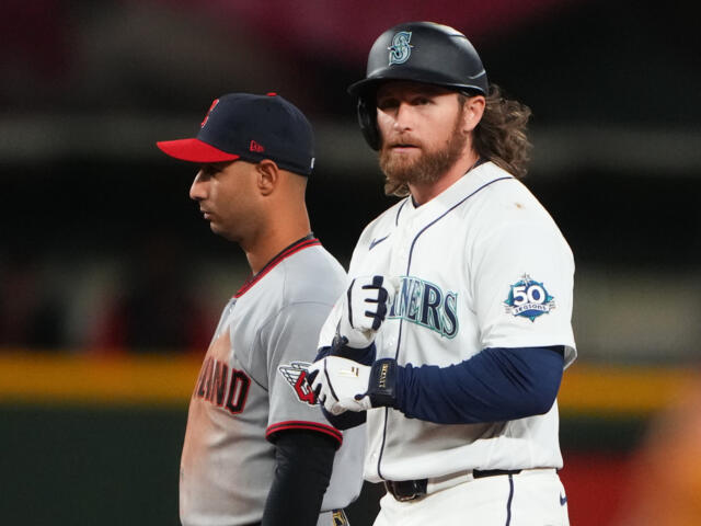 Seattle Mariners batter in white uniform with helmet and 50 Seasons patch stands at the plate, while a Cleveland pitcher/infielder in gray with a red cap lingers behind him.