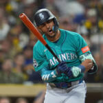 Mariners batter mid-swing in teal uniform with orange bat during a baseball game, crowd in background