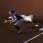Baseball infield action: fielder in blue reaches for a ground ball as a baserunner slides toward the base beside him.