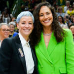 Two women stand together and smile at a crowded event, the left woman wearing a black blazer and white shirt with a decorative brooch, the right in a bright green blazer.