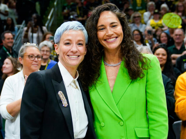 Two women stand together and smile at a crowded event, the left woman wearing a black blazer and white shirt with a decorative brooch, the right in a bright green blazer.