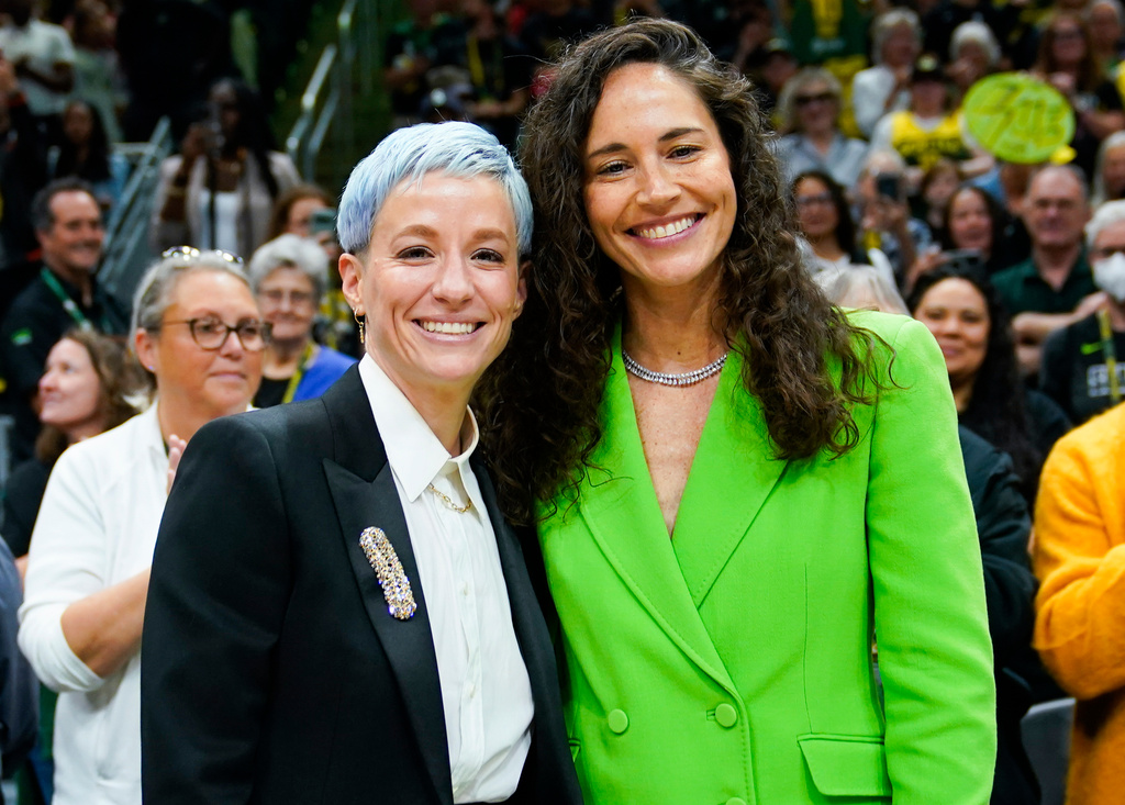Two women stand together and smile at a crowded event, the left woman wearing a black blazer and white shirt with a decorative brooch, the right in a bright green blazer.