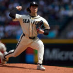 Baseball pitcher in a Seattle Mariners uniform winding up to throw on the dirt mound with a blurred crowd behind him.
