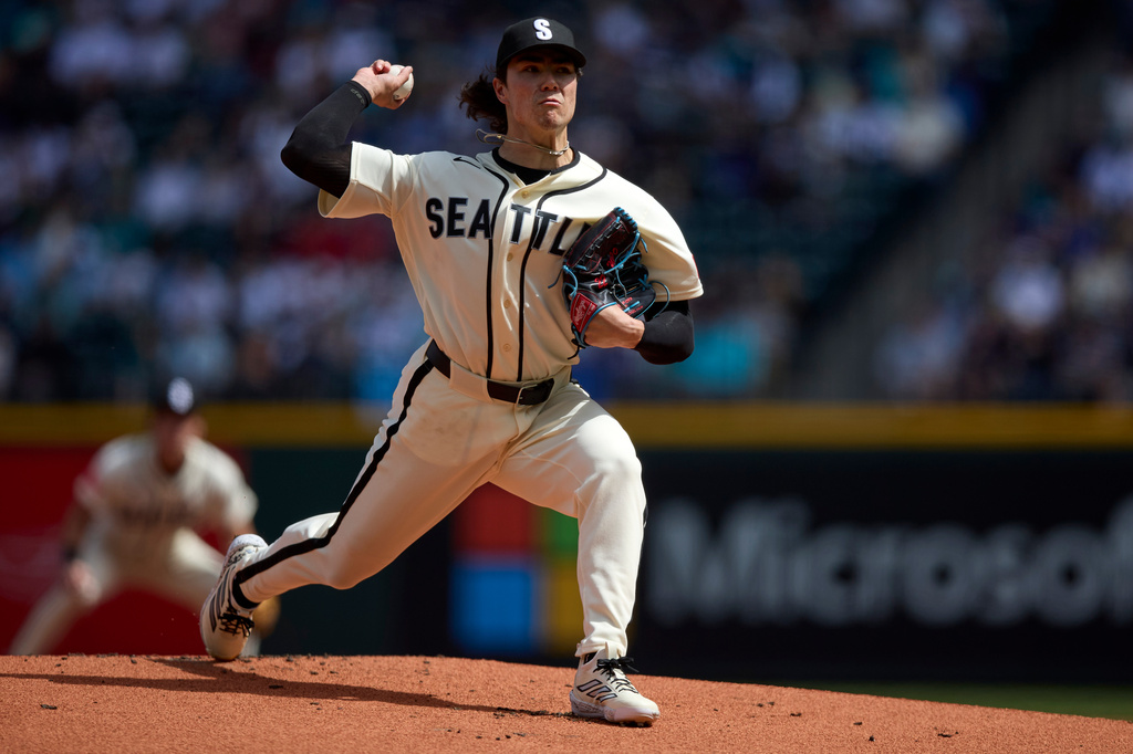 Baseball pitcher in a Seattle Mariners uniform winding up to throw on the dirt mound with a blurred crowd behind him.