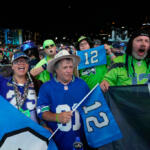 Group of Seattle Seahawks fans in blue and green jerseys cheering at a nighttime stadium, holding banners and flags.