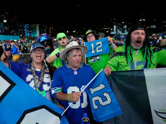 Group of Seattle Seahawks fans in blue and green jerseys cheering at a nighttime stadium, holding banners and flags.