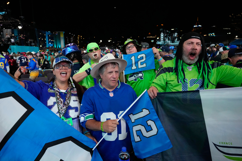 Group of Seattle Seahawks fans in blue and green jerseys cheering at a nighttime stadium, holding banners and flags.