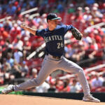 Baseball pitcher in a Seattle uniform delivering a pitch on the mound with a crowd watching.