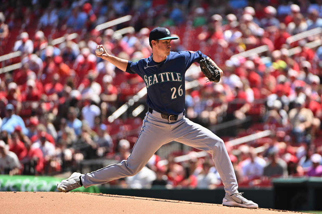 Baseball pitcher in a Seattle uniform delivering a pitch on the mound with a crowd watching.