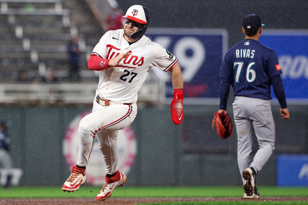 Twins player wearing #27 in a white uniform with red accents runs on a wet infield as a blue-team fielder (#76, Rivas) watches in the rain.
