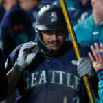 Seattle Mariners player in a dark-blue uniform holds a golden bat, ready to swing, as teammates give a high-five nearby.