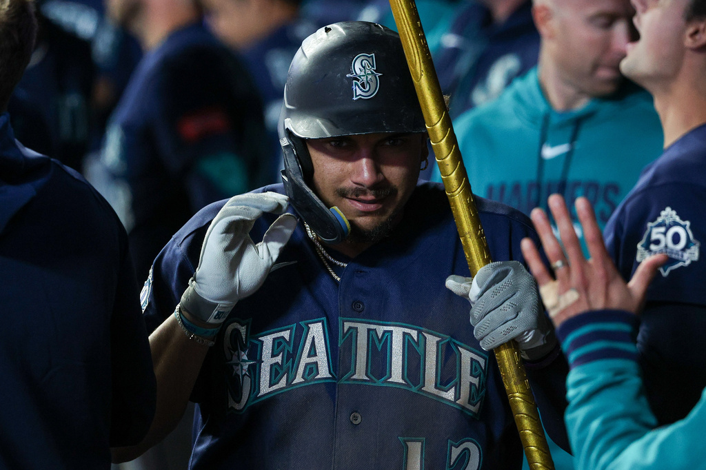 Seattle Mariners player in a dark-blue uniform holds a golden bat, ready to swing, as teammates give a high-five nearby.
