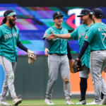 Seattle Mariners players in teal uniforms celebrate on the field, high-fiving and smiling together.