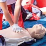 Person in red pants practicing chest compressions on a CPR manikin during a training drill with gloved hands.