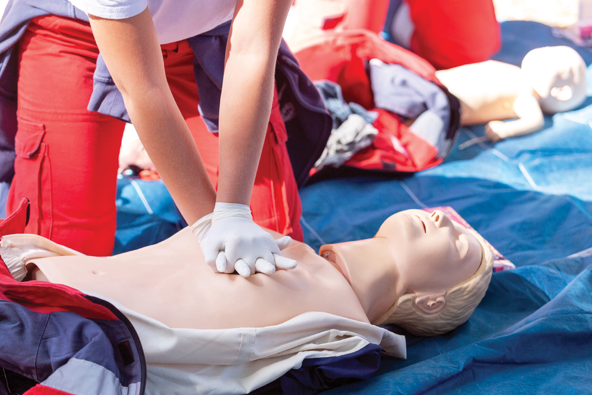 Person in red pants practicing chest compressions on a CPR manikin during a training drill with gloved hands.