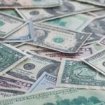 Pile of assorted U.S. dollar bills scattered and overlapping on a flat surface (including $1, $5, $10, and $100 notes).