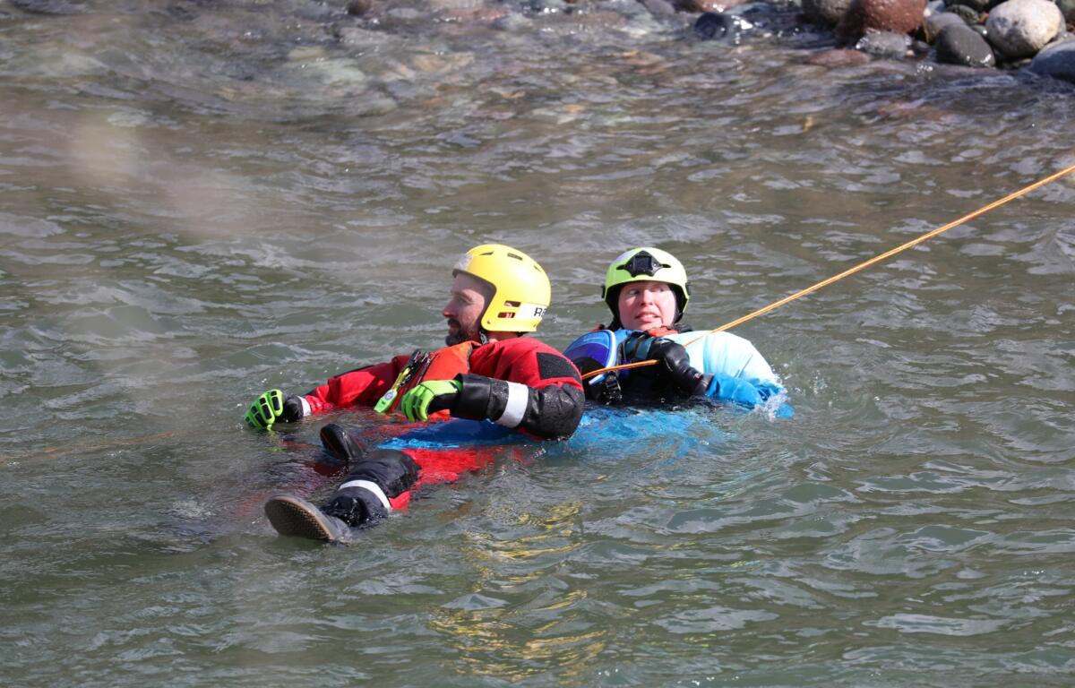 Two people wearing helmets and life jackets float together in a river, gripping a safety rope.