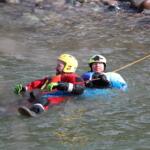 Two people wearing helmets and life jackets float together in a river, gripping a safety rope.