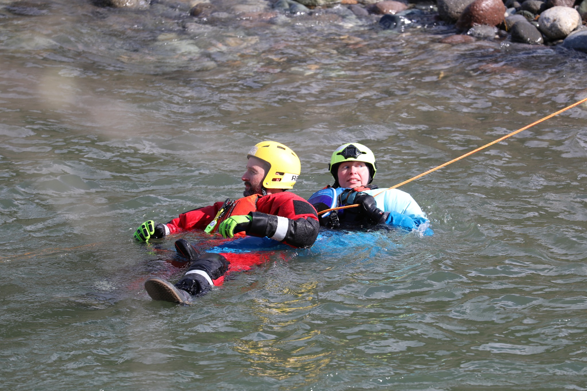 Two people wearing helmets and life jackets float together in a river, gripping a safety rope.