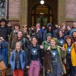 Group of diverse students posing on campus steps, fists raised in solidarity.