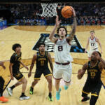 Gonzaga player in white jersey #8 leaps toward the basket for a layup as defenders in black Kennesaw State uniforms swarm around him with the crowd in the stands behind.