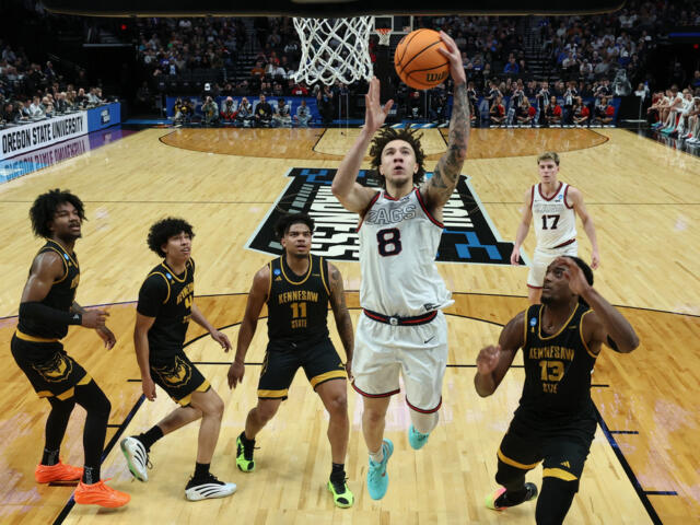 Gonzaga player in white jersey #8 leaps toward the basket for a layup as defenders in black Kennesaw State uniforms swarm around him with the crowd in the stands behind.