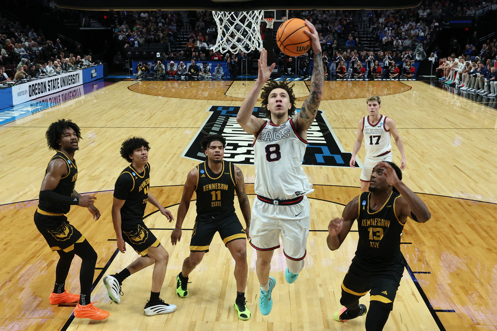 Gonzaga player in white jersey #8 leaps toward the basket for a layup as defenders in black Kennesaw State uniforms swarm around him with the crowd in the stands behind.