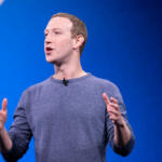 Man in a blue sweater giving a presentation on stage, gesturing with his hands.