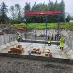 Construction site with a large concrete basin, workers in high‑visibility clothing, and a red beam being lowered overhead by a crane.