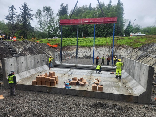 Construction site with a large concrete basin, workers in high‑visibility clothing, and a red beam being lowered overhead by a crane.