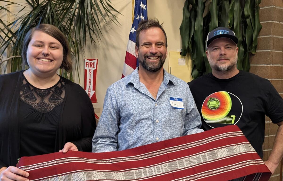 Three people smile at the camera indoors, holding a red woven textile together with an American flag in the background to the center.