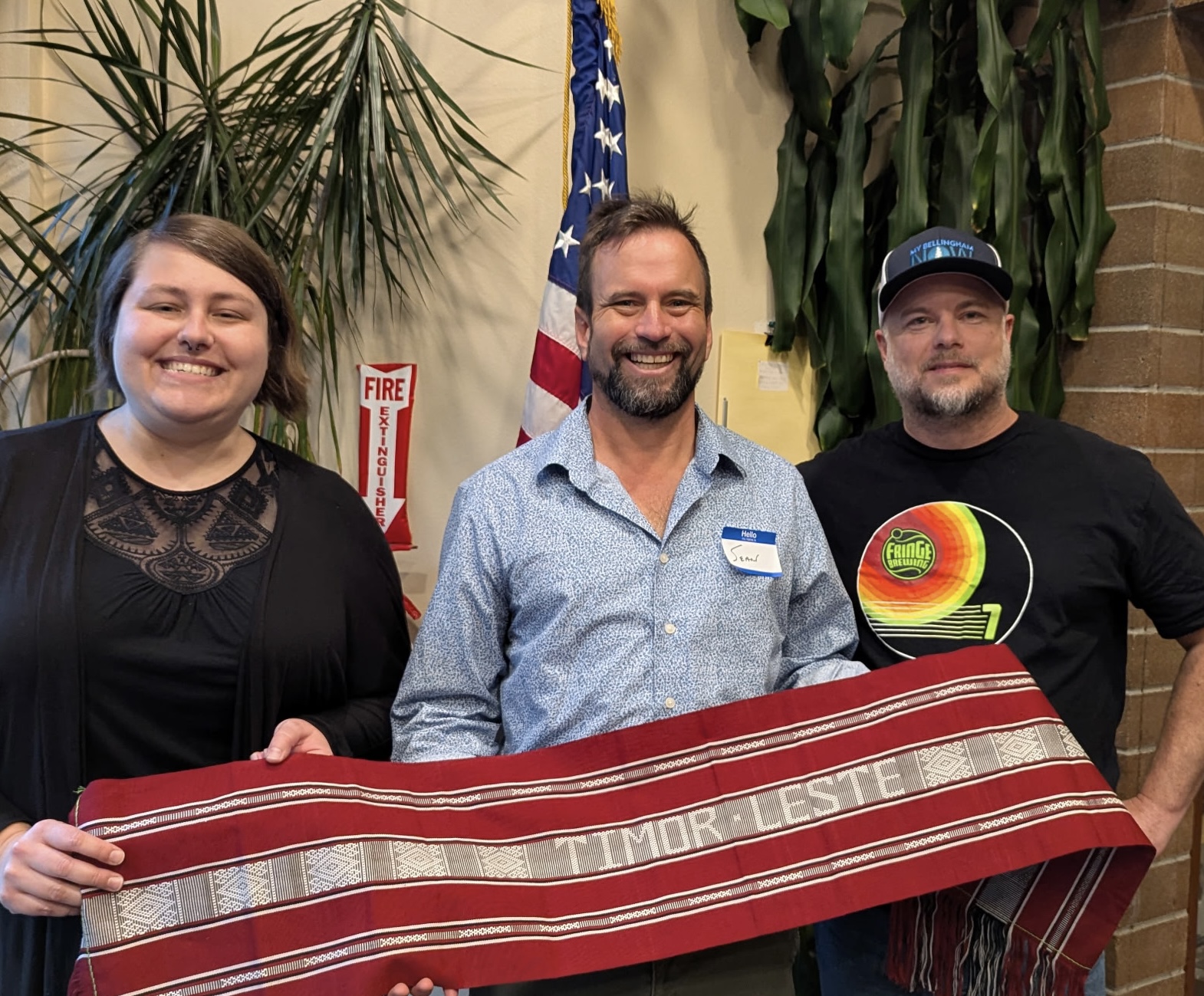 Three people smile at the camera indoors, holding a red woven textile together with an American flag in the background to the center.