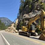 Yellow excavator blocking a mountain road after a rock slide, with a worker in orange near the guardrail on the left and rocky cliff on the right