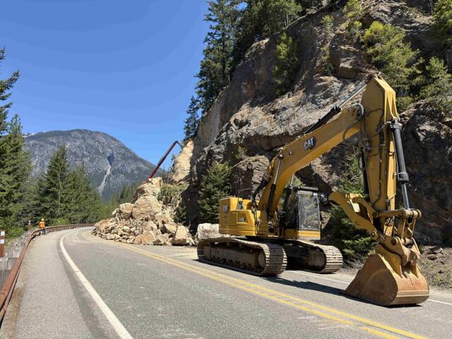 Yellow excavator blocking a mountain road after a rock slide, with a worker in orange near the guardrail on the left and rocky cliff on the right