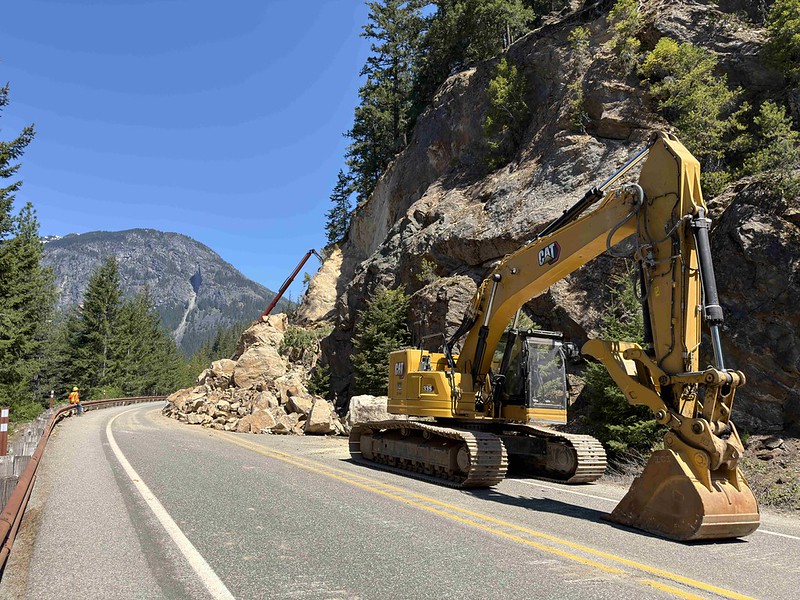 Yellow excavator blocking a mountain road after a rock slide, with a worker in orange near the guardrail on the left and rocky cliff on the right