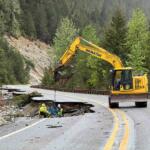 Yellow excavator on a mountain road repairing a sinkhole with two workers nearby; forested hills in the background.