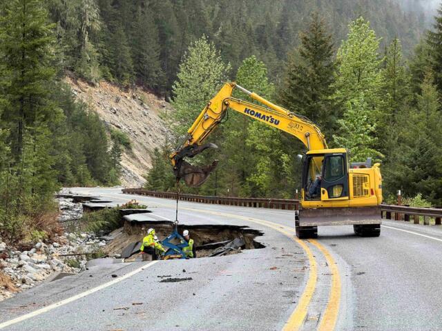 Yellow excavator on a mountain road repairing a sinkhole with two workers nearby; forested hills in the background.