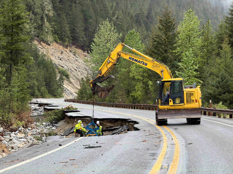 Yellow excavator on a mountain road repairing a sinkhole with two workers nearby; forested hills in the background.