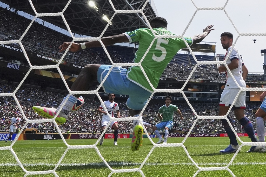 Goalkeeper in a green jersey with number 5 dives toward the ball as it hits or crosses the net; a white-clad defender stands nearby in a packed stadium.