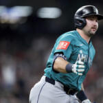 Baseball player in teal Mariners uniform running on the field with a blurred crowd in the background, helmet on and gloves on.