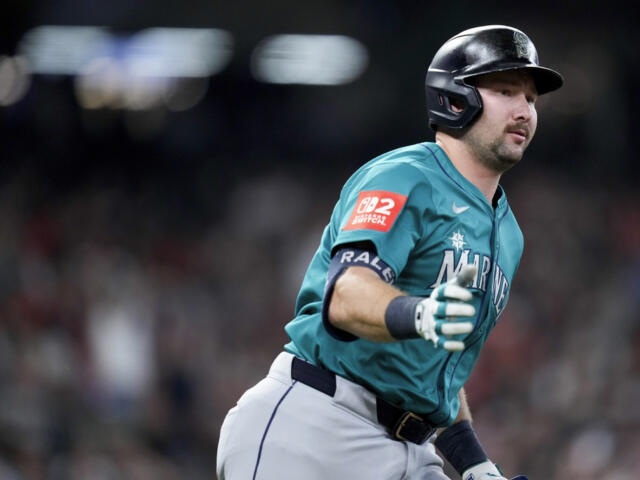 Baseball player in teal Mariners uniform running on the field with a blurred crowd in the background, helmet on and gloves on.