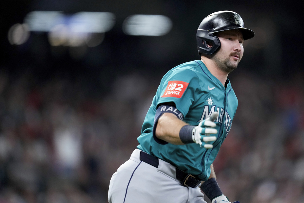 Baseball player in teal Mariners uniform running on the field with a blurred crowd in the background, helmet on and gloves on.