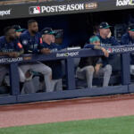 Seattle Mariners players in the dugout during a postseason game, leaning on the railing and watching the field, with Postseason 2025 and loanDepot signage visible.
